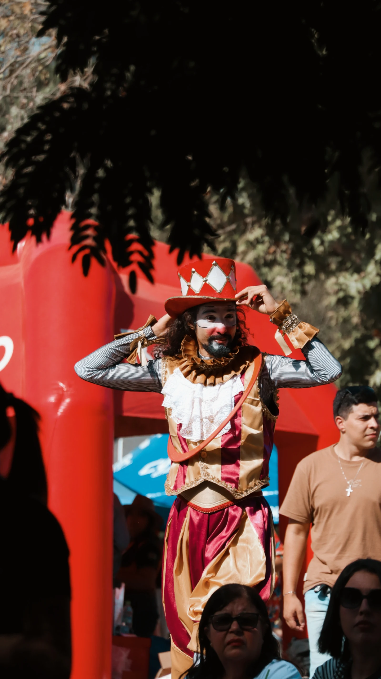 Artista caracterizado com maquiagem de palhaço e figurino vermelho e dourado, ajustando o chapéu durante um evento ao ar livre.